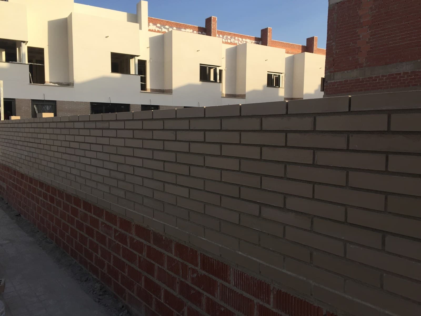 Muro de ladrillo caravista gris con las juntas a la vista, levantado sobre unas hiladas de ladrillo rojo, delante de una fila de adosados en obra