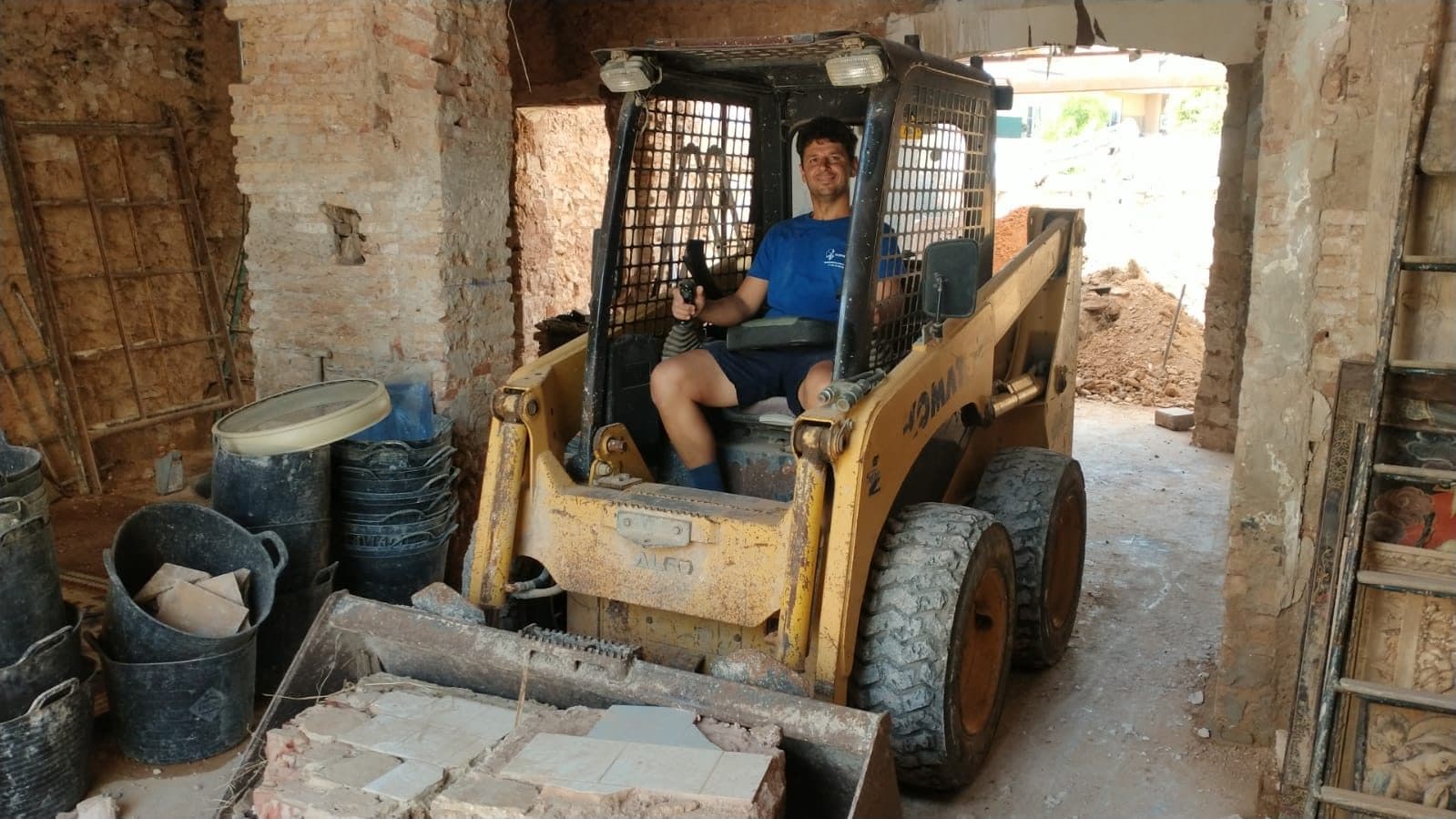 Uno del equipo a los mandos de una minicargadora con escombro en la pala, en una casa en rehabilitación con los muros de ladrillo a la vista